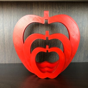 May include: Three red wooden apples nested inside each other. The apples are all the same shape, but different sizes. The largest apple is at the bottom, and the smallest apple is at the top.