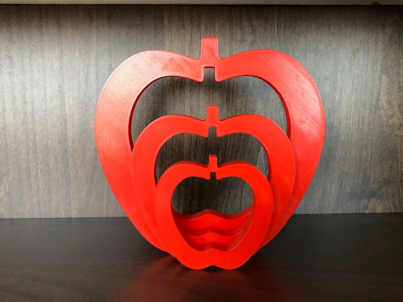 May include: Three red wooden apples nested inside each other. The apples are all the same shape, but different sizes. The largest apple is at the bottom, and the smallest apple is at the top.