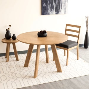 May include: A round wooden dining table with four legs and a white rug underneath. A chair with a grey cushion is pulled up to the table. There is a small wooden side table with a black vase and a small vase with a single flower.