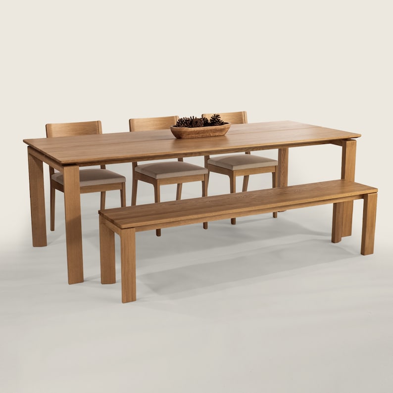 May include: A wooden dining table set with four chairs and a bench. The table is made of light brown wood and has a rectangular shape. The chairs are also made of light brown wood and have a simple design. The bench is made of the same wood and has a rectangular shape. The table is set with a bowl of pine cones in the center.