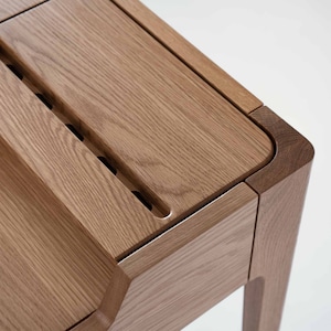 May include: Close-up of a wooden desk with a unique design featuring a built-in drawer and a groove for organizing items. The desk is made of light brown wood with a natural grain pattern.