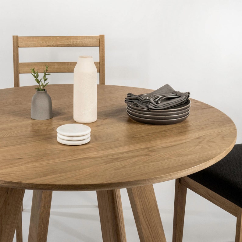 May include: A round wooden table with a light brown finish. The table has a simple design with four legs and a smooth surface. There are two white coasters and a white ceramic vase with a single green stem on the table. A stack of grey napkins and plates are on the table.