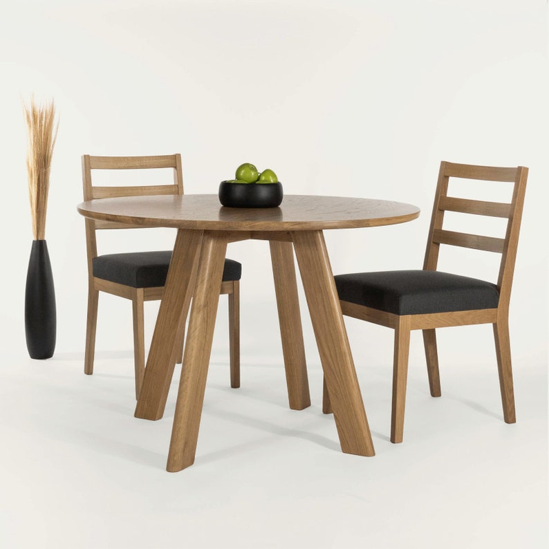 May include: A round wooden dining table with a natural wood finish and three legs. Two chairs with black fabric seats and wooden frames are placed around the table. A small bowl of green apples sits on the table.
