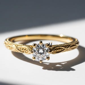Puede incluir: Un anillo de compromiso dorado con un diamante redondo y transparente en el centro. La banda presenta un diseño intrincado y grabado. El anillo está sobre un fondo blanco, con sombras.
