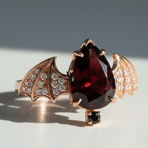 May include: Rose gold ring featuring a pear-shaped, deep red gemstone. The ring has bat wing designs on either side, encrusted with small, clear gemstones. A small black gemstone hangs below the main stone.