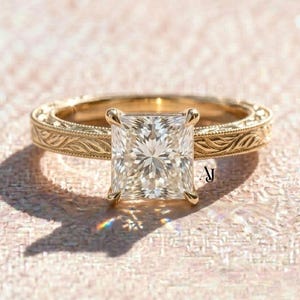 May include: A gold engagement ring with a square-cut diamond. The ring features an engraved band with a floral pattern. The diamond is held in place by four prongs. The ring is set against a soft pink background.