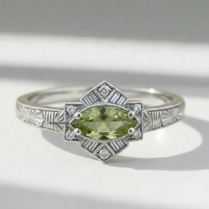 Può includere: Un anello d'argento con una pietra preziosa verde chiaro a taglio marquise al centro, circondata da pietre chiare più piccole e dettagli baguette. La fascia ha un motivo inciso. Un bellissimo gioiello.