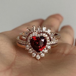 May include: A heart-shaped red gemstone ring with a halo of smaller diamonds set in a rose gold band. The ring is surrounded by a delicate band of diamonds.