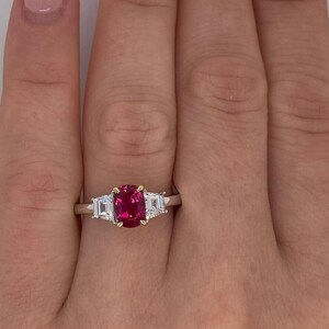 May include: A gold and silver ring featuring a large, oval-cut, bright red gemstone as the center stone, flanked by two smaller, rectangular, clear gemstones. The ring is set on a hand with a light skin tone.