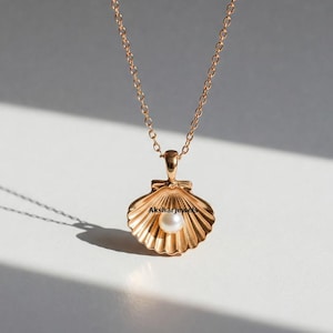 Può includere: Una collana con pendente a conchiglia color oro con una catena delicata. Il pendente presenta un design a conchiglia dettagliato con una piccola perla rotonda al centro. La collana è esposta su una superficie bianca con ombre.