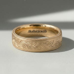 Puede incluir: Un anillo de color dorado con un patrón de hojas grabado alrededor de la banda. El anillo tiene un borde texturizado y el texto "Aksharjewels" es visible en el anillo.