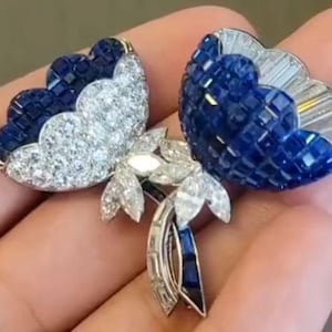 Puede incluir: Un par de pendientes con forma de mariposa. Cada pendiente presenta un diseño de ala con piedras preciosas azules y transparentes. Las alas están detalladas con piedras redondas y de talla baguette, creando un efecto brillante. Los pendientes están engarzados en un metal plateado.