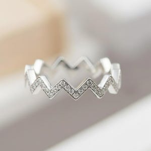 Könnte beinhalten: Ein silberfarbener Ring mit Zickzack-Muster. Der Ring ist mit kleinen, klaren Edelsteinen entlang des Zickzack-Musters besetzt. Der Ring ist vor einem weichen, verschwommenen Hintergrund platziert.