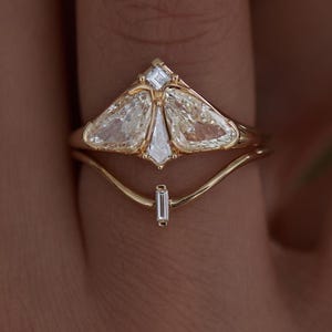Può includere: Un set di anelli d'oro con un design unico con più pietre preziose chiare e sfaccettate. L'anello superiore ha una forma triangolare con due pietre a forma di pera e una pietra quadrata più piccola. Un secondo anello con una pietra taglio baguette si trova sotto.