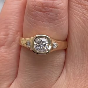 May include: A gold ring featuring a large, round, bezel-set diamond with two smaller diamonds on the sides.