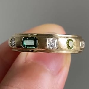 Puede incluir: Un anillo de banda de oro con cinco piedras preciosas. El anillo tiene una esmeralda verde, un diamante transparente, un peridoto verde y dos diamantes transparentes más pequeños.