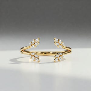 Könnte beinhalten: Ein goldfarbener Ring mit Blattdesign. Der Ring hat kleine, klare Edelsteine, die in blattartigen Clustern angeordnet sind. Das Band des Rings ist offen, mit den Blattclustern an jedem Ende. Der Markenname "Aksharjewels" ist auf dem Ring sichtbar.