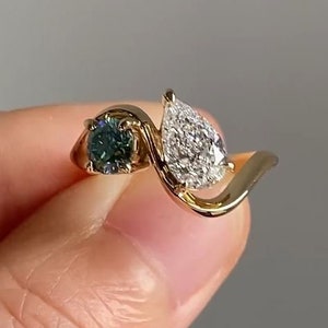 Puede incluir: Un anillo de oro con un diamante en forma de pera y una piedra preciosa redonda verde. El anillo tiene un diseño único y retorcido.