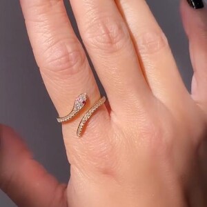 May include: A gold-coloured snake ring featuring a red gemstone at the head and small clear gemstones along the body. The ring wraps around the finger, creating a stylish and unique design.
