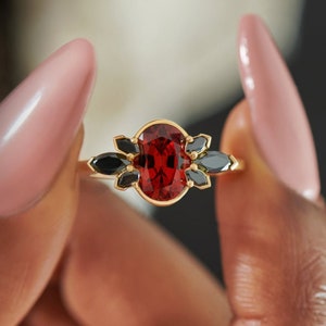 Könnte beinhalten: Ein goldener Ring mit einem großen roten ovalen Edelstein, der von kleineren schwarzen Edelsteinen umgeben ist.