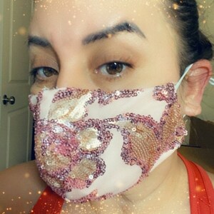 May include: A close-up of a person wearing a white face mask with pink and gold sequins. The mask has a floral pattern and is made of a sheer fabric.