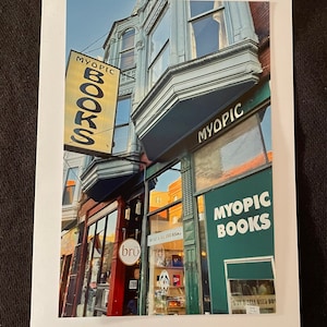 May include: A shopfront with a green awning that reads "Myopic Books" in white lettering. The shopfront is on a corner and has a yellow sign that reads "Myopic Books" in black lettering. The sign is hanging from the building above the shopfront.