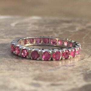 Può includere: Un anello d'argento con una fila continua di pietre preziose rosa. Le pietre sono rotonde e ravvicinate, creando una fascia di colore vivace. L'anello è esposto su una superficie neutra e strutturata.