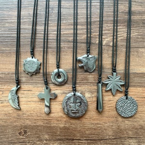 May include: Nine black obsidian pendants on black cords, each with a different design: a crescent moon, a sun face, a cross, a lion's head, a wolf's head, a circle with a hole in the center, a pointed obelisk, a marijuana leaf, and a textured circle.