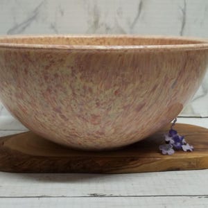 May include: A large, round mixing bowl with a speckled, marbled pattern in shades of peach, cream, and light brown. The bowl has a smooth, curved rim and a deep basin, resting on a natural, oval-shaped wooden board with a visible grain.