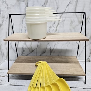 May include: A set of white plastic measuring cups stacked vertically on a two-tiered wooden shelf with a black metal frame, accompanied by a set of yellow plastic measuring spoons fanned out on the lower shelf surface.