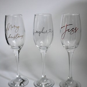 May include: Three clear champagne flutes with text. One flute has "Merry Christmas" in gold script, another has "sophie" with a heart, and the third has "Jess" in red script. The flutes are on a white background.