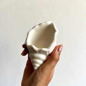 以下が含まれることがあります： 白いセラミック製の貝殻型プランター。プランターは、テクスチャ加工された螺旋状のベースと、滑らかで開いた上部を備えています。貝殻は、無地の白い背景に対して手で持たれています。プランターは、小さな植物や装飾品を入れるように設計されています。