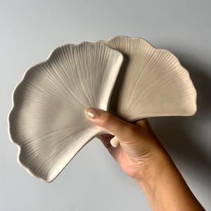 Op de afbeelding: Twee beige, waaiervormige keramische schalen. De schalen hebben een textuur, geaderd ontwerp, dat lijkt op een ginkgoblader. De schalen worden tegen een lichtgrijze achtergrond gehouden.
