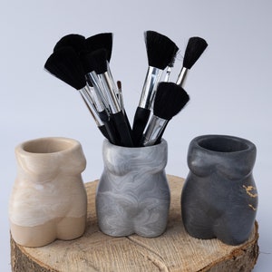 Puede incluir: Tres contenedores de cerámica decorativos con forma de torso humano, en beige, gris y negro, con un montón de pinceles de maquillaje negros en el contenedor gris. Los contenedores están sobre una rodaja de tronco de madera.
