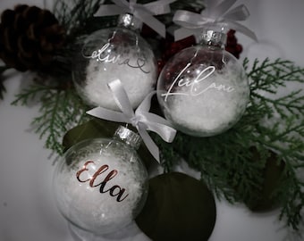 May include: Three clear glass Christmas baubles with white fluffy filling and personalized names in cursive script: "Olivia", "Leilani", and "Ella". Each bauble is tied with a silver ribbon. The baubles are arranged with evergreen branches and a pinecone.