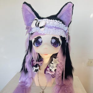 Pode incluir: Um capacete de cosplay com tema de gato com olhos grandes e expressivos e cabelo longo e esvoaçante lavanda e preto. O capacete apresenta orelhas grandes e pontudas, uma faixa de renda branca e laços decorativos. Um colar de cruz de prata pende.