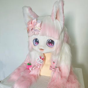 Pode incluir: Uma cabeça de fantasia de coelho de pelúcia branca e rosa com olhos grandes e expressivos e orelhas longas e fofas. A fantasia apresenta um laço rosa e detalhes de morango, com longas extensões de pele rosa.