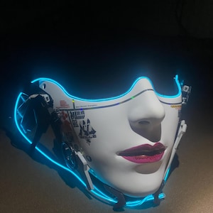 May include: A white face mask with a feminine design, featuring pink lips and a blue neon outline. The mask has Japanese text and mechanical details, creating a futuristic aesthetic.