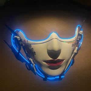 May include: A white face mask with a stylized female face, red lips, and a blue neon outline. The mask has mechanical details on the sides and text on the surface. The mask is designed with a futuristic aesthetic.