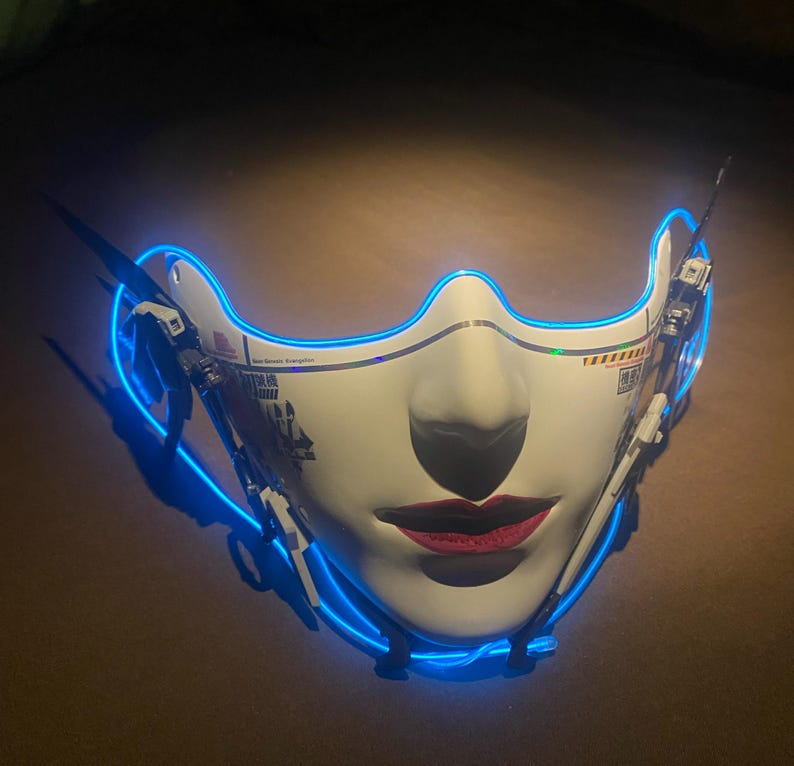 May include: A white face mask with a stylized female face, red lips, and a blue neon outline. The mask has mechanical details on the sides and text on the surface. The mask is designed with a futuristic aesthetic.