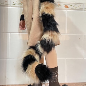 May include: A faux fur tail and leg warmers in black, tan, and cream. The tail is long and fluffy, while the leg warmers are shorter and more compact. The accessories are worn with black platform sandals and brown socks.