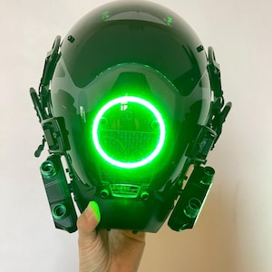 Può includere: Un casco futuristico verde scuro con una luce circolare verde brillante al centro. Il casco ha diversi attacchi e una visiera trasparente. Il design suggerisce un tema fantascientifico o cosplay.