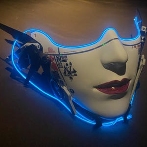 May include: A white futuristic mask with a neon blue outline, depicting a stylized face. The mask features red lips and black accents, with Japanese text and mechanical details. The design evokes a cyberpunk or science fiction aesthetic.