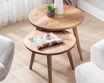 May include: Two round wooden nesting tables with a natural wood finish. The top table has a vase with green plants and a small potted plant. The bottom table has a magazine on top.