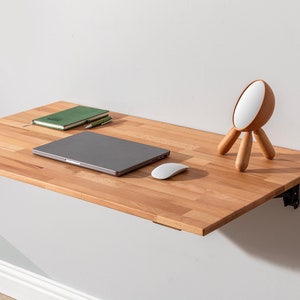 May include: A light wood folding wall desk with a laptop, mouse, and a small wooden lamp on top. The desk is mounted to a white wall.