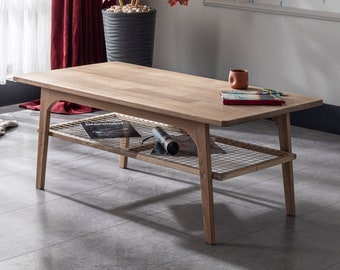 May include: A light oak coffee table with a lower wire mesh shelf. The table has a rectangular top and four angled legs. A magazine and a small cup are on the table's surface. The table's length is approximately 120 cm.