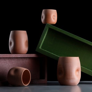 Può includere: Quattro tazze in terracotta di colore terracotta con una superficie testurizzata sono disposte su una piattaforma di legno. Una tazza è in equilibrio su una scatola di legno verde, che è inclinata di lato. Le tazze sono tutte della stessa dimensione e forma, e hanno un aspetto unico e organico.