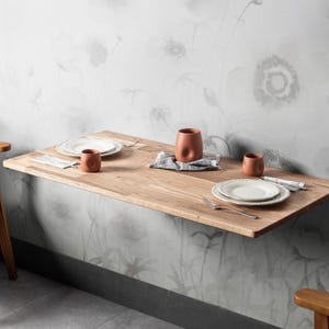 May include: A wall-mounted wooden table with a natural finish, set with white plates, terracotta-colored cups, and a small gray cloth. The table is against a floral wallpaper background, creating a minimalist dining setting.