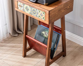May include: A wooden record player stand with a drawer featuring a colorful tile design. The stand has a built-in magazine rack with a brown leather flap and a shelf for storing records.