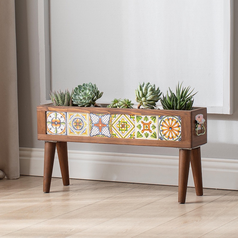 May include: A wooden planter box with a decorative tile design in shades of blue, yellow, and green. The planter is on legs and has a small metal handle on the side. It is filled with succulents.